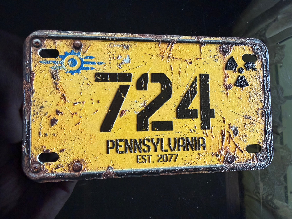 A distressed yellow novelty license plate with black lettering reading '724 PENNSYLVANIA EST. 2077', featuring a blue circular logo on the left and a black radiation symbol on the right, held against a dark background.