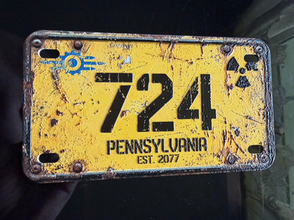 A distressed yellow novelty license plate with black lettering reading '724 PENNSYLVANIA EST. 2077', featuring a blue circular logo on the left and a black radiation symbol on the right, held against a dark background.