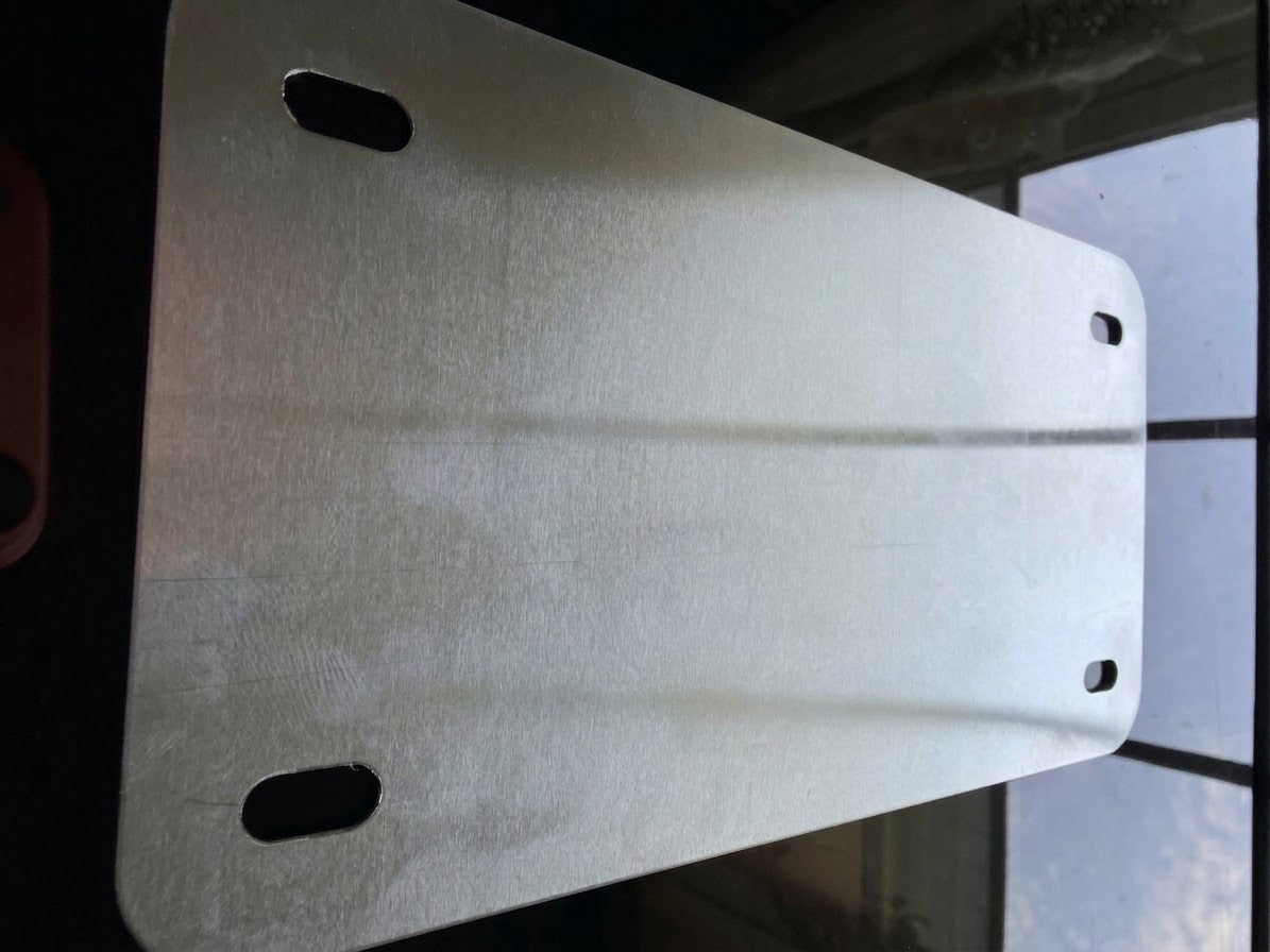 A brushed silver metal mounting plate with rounded corners and four pre-drilled holes, shown against a dark background with a window pane visible on the right.