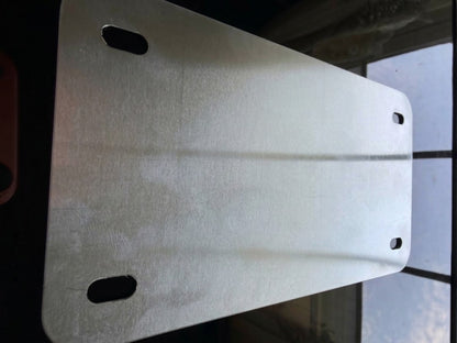A brushed silver metal mounting plate with rounded corners and four pre-drilled holes, shown against a dark background with a window pane visible on the right.